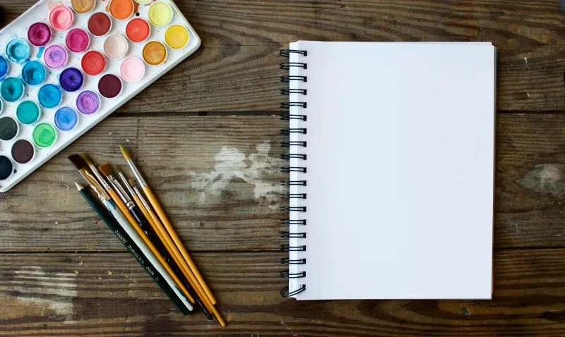 Paint brushes beside a blank spiral sketchbook and a watercolour palette on a wooden table