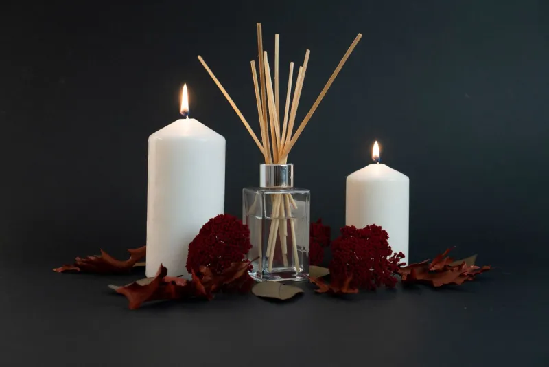 A glass reed diffuser between two lit white pillar candles with dried red flowers and autumn leaves on a dark background