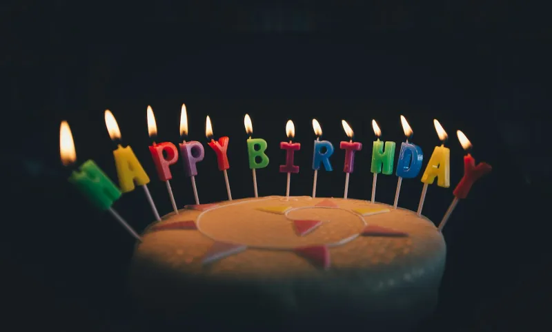 A round iced cake with lit Happy Birthday letter candles against a dark background