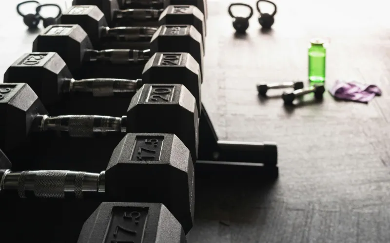 A rack of black hex dumbbells with kettlebells and a water bottle on a gym floor