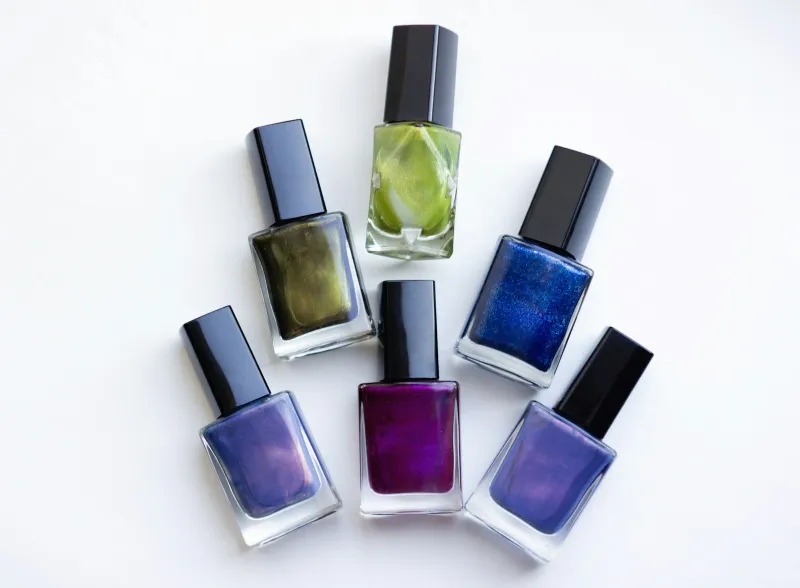 Six bottles of purple, blue and green nail polish arranged on a white surface