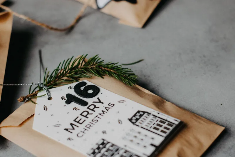 A brown paper advent calendar bag with a number 16 Merry Christmas tag and a sprig of fir on a grey surface
