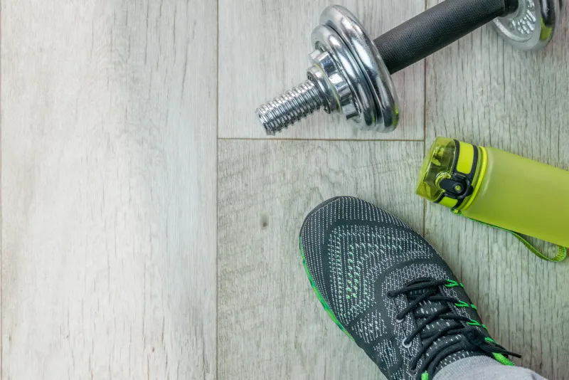 A trainer, dumbbell and green water bottle on a light wooden floor