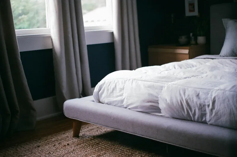 Bed with a thick white duvet beside a window with grey curtains