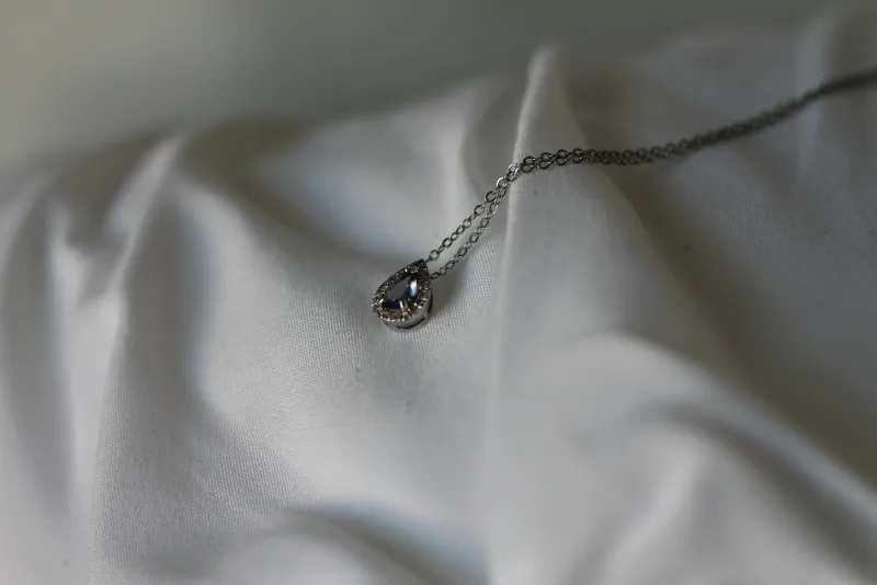 A silver pendant necklace with a small teardrop stone resting on folded white fabric