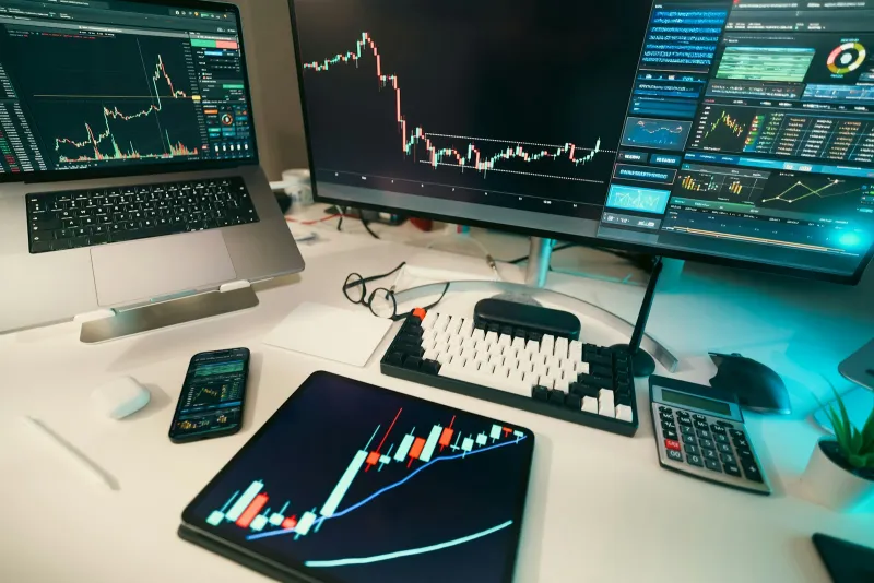 A desk with a laptop, monitor, tablet and phone all showing candlestick trading charts