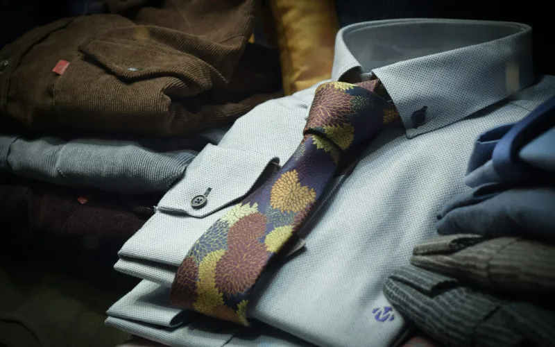 A patterned tie on a folded pale blue dress shirt beside a stack of jackets