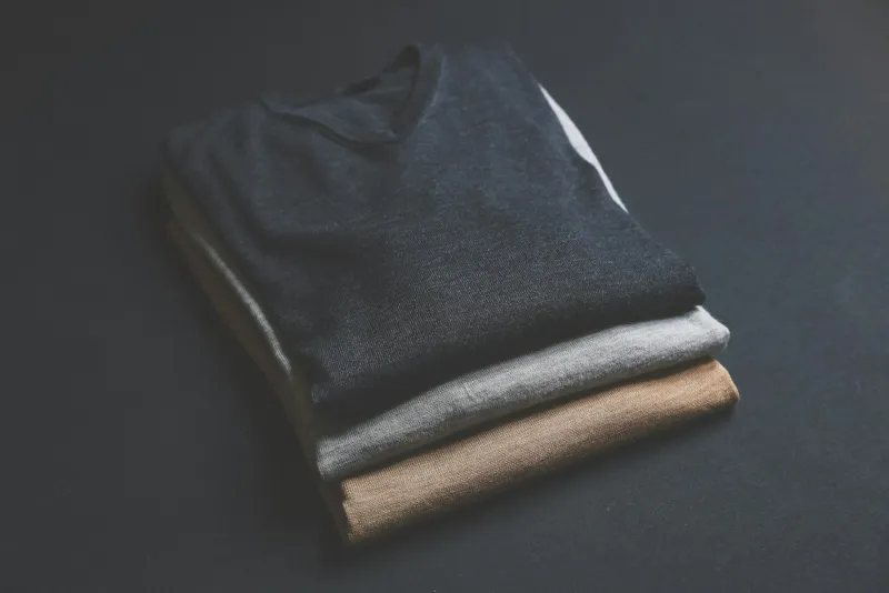 Three folded V-neck jumpers in navy, grey and camel stacked on a dark surface