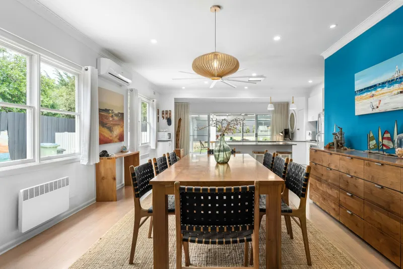 A bright dining room with a long wooden table, woven black chairs and a wooden sideboard