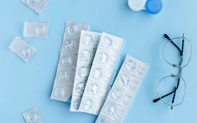 Blister packs of contact lenses, a lens case and a pair of glasses on a pale blue surface