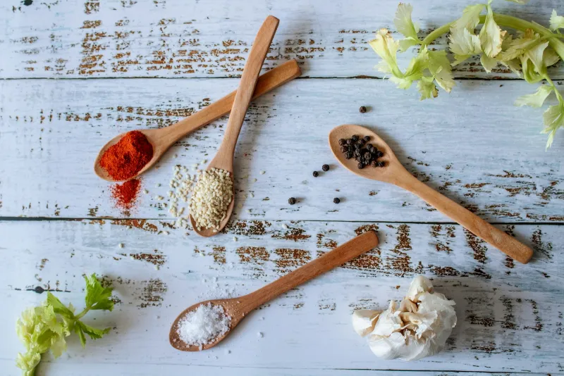 Wooden spoons of sea salt flakes, black peppercorns, paprika and sesame seeds on a white wooden table