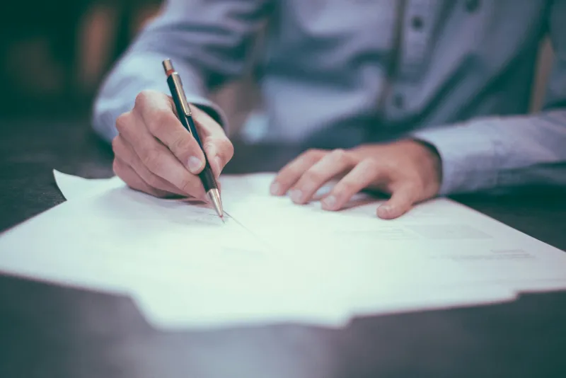 A person in a blue shirt signing a paper document with a pen at a desk