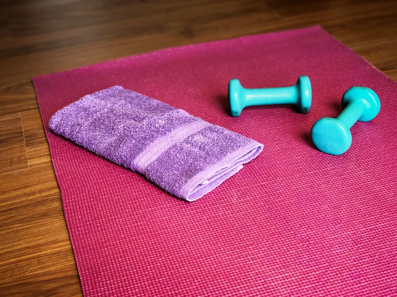 Pink exercise mat with a folded towel and two small dumbbells on a wooden floor