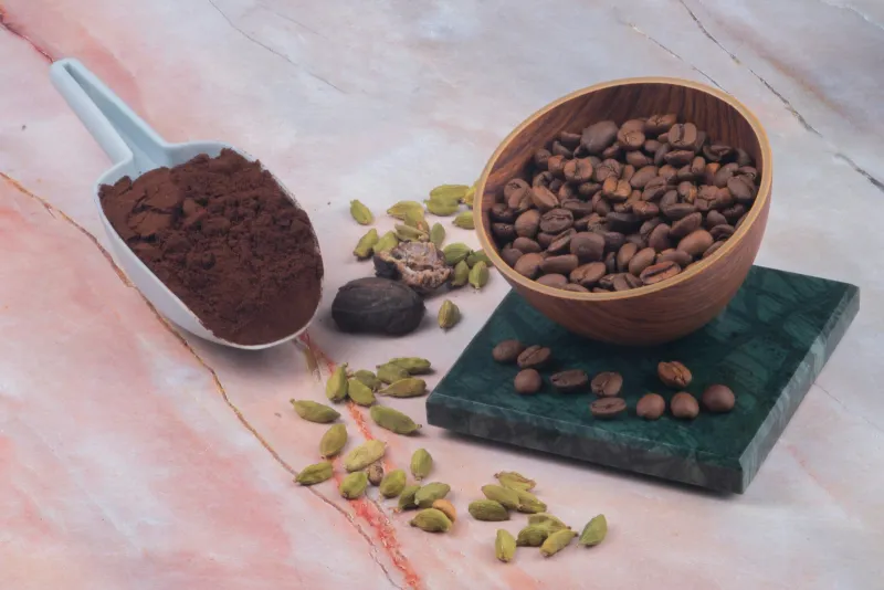 A wooden bowl of coffee beans beside a scoop of cocoa powder and cardamom pods on marble