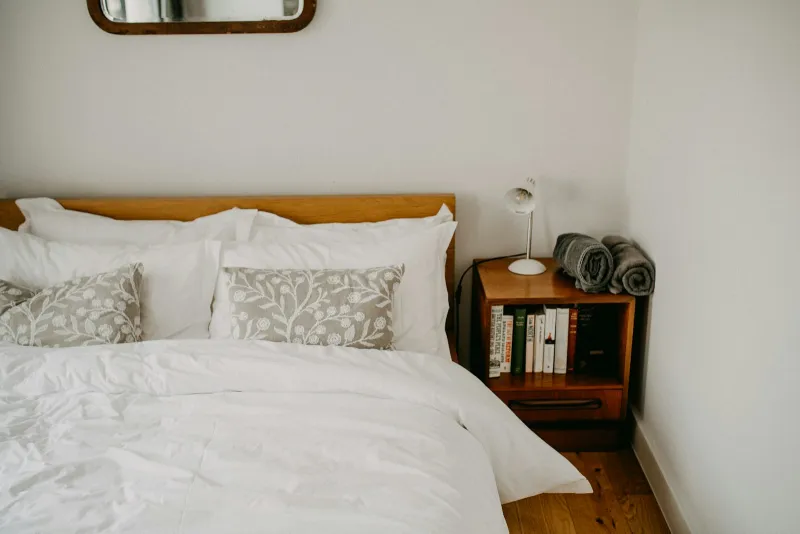 A neatly made bed with white linen and patterned cushions beside a wooden bedside table with books and a lamp