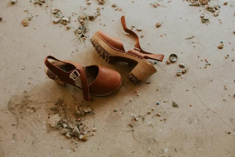 A pair of tan leather clogs with ankle straps lying on a dusty concrete floor