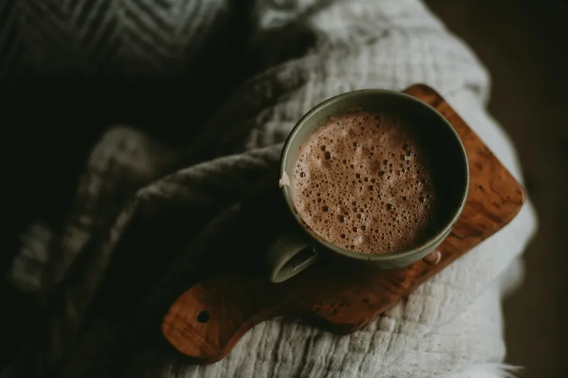 Cup of frothy hot chocolate on a wooden board resting on a knitted blanket