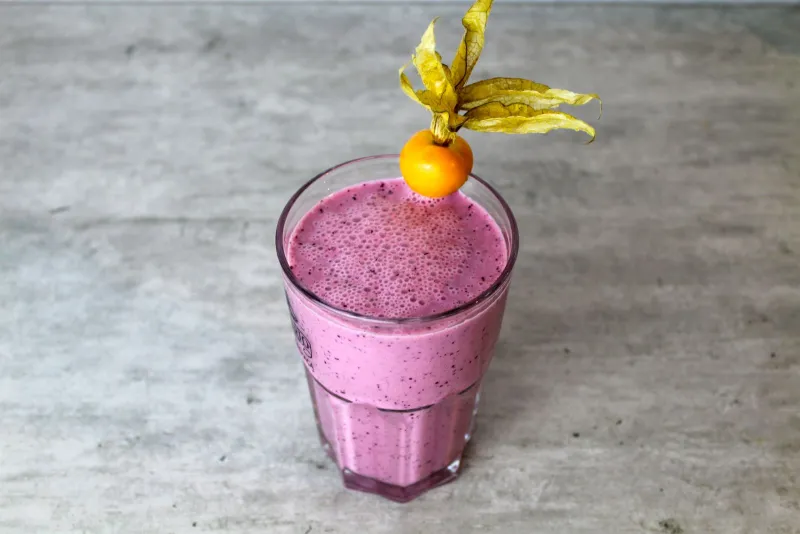 A pink berry smoothie in a glass topped with a physalis on a grey worktop
