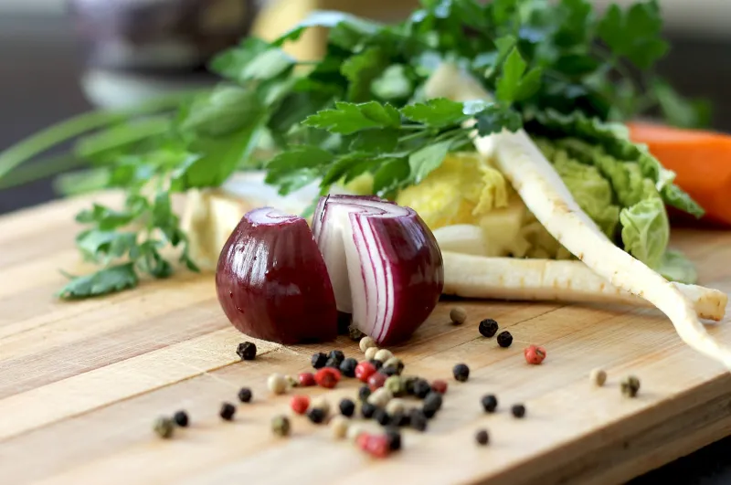 A halved red onion, parsley, parsnip and peppercorns on a wooden chopping board