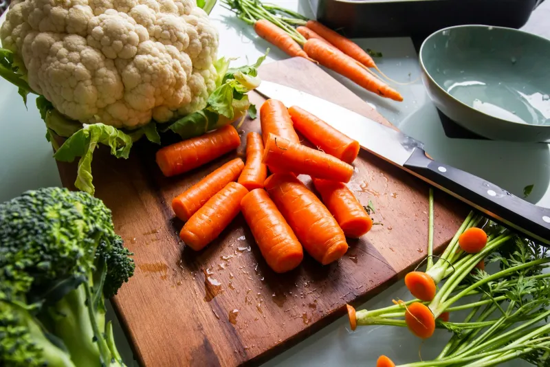 Chopped carrots and a kitchen knife on a wooden chopping board, with a cauliflower and broccoli beside them