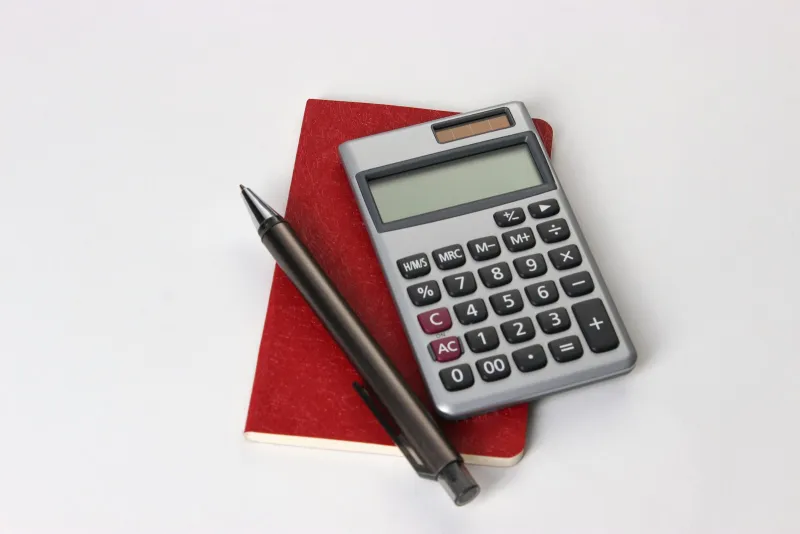 A silver pocket calculator and a black pen resting on a red notebook against a white background