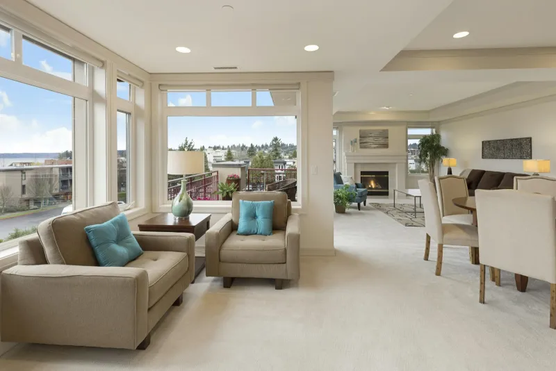 A bright, tidy open-plan living room with beige armchairs, a dining table and a fireplace
