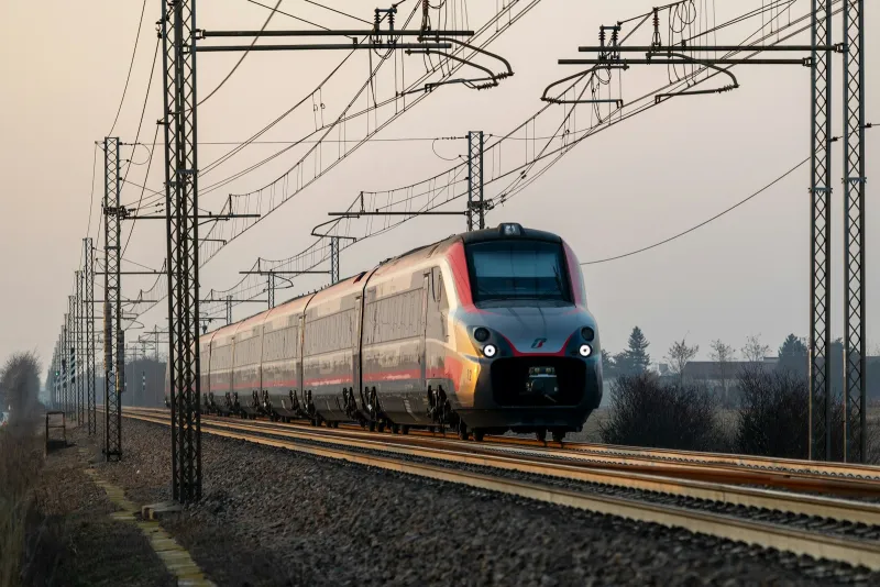 A high-speed train running along tracks under overhead power lines in hazy golden light