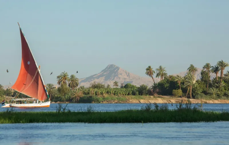 A traditional felucca with a red sail on the Nile at Luxor, with palms and desert hills behind