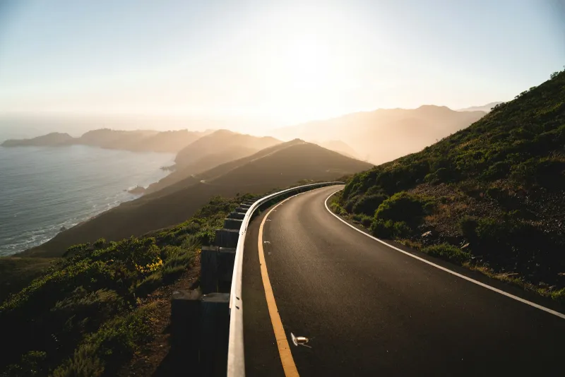 An empty coastal road curving along green hills above the sea at sunset