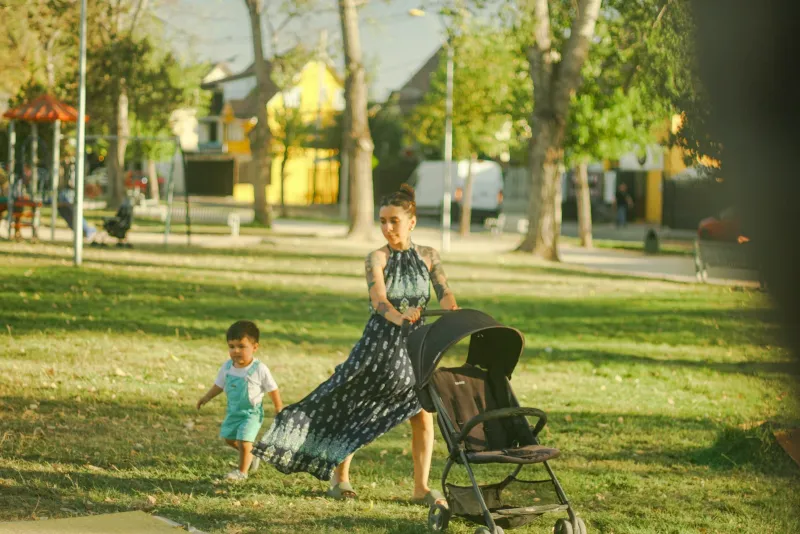Woman pushing a pushchair through a sunny park with a toddler walking alongside