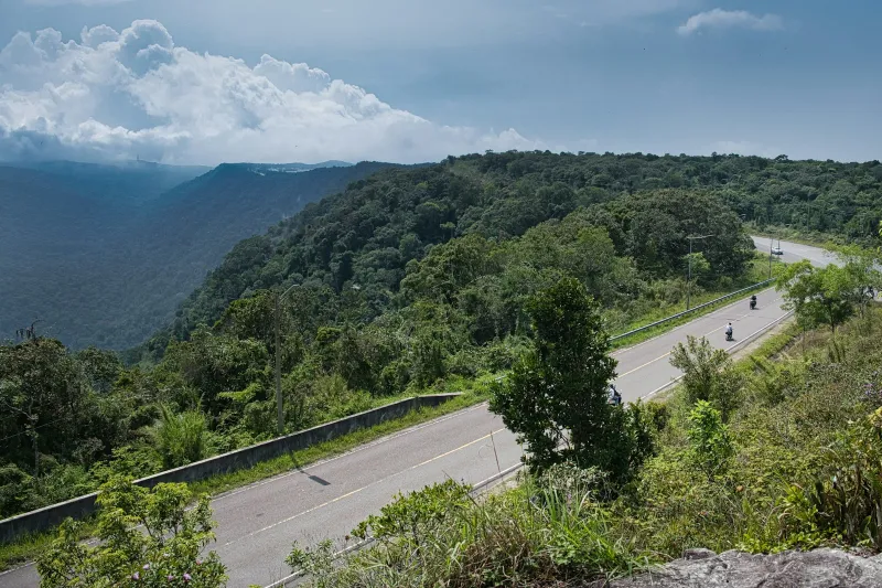 Motorcyclists riding a road along a forested hillside under a cloudy sky