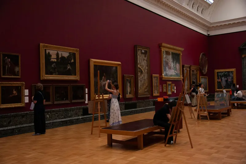 Visitors looking at paintings in gilt frames on a deep red gallery wall