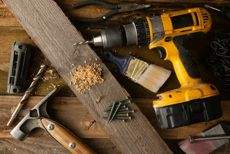 A cordless drill, hammer, pliers, paintbrush, drill bit and screws on a rough wooden workbench