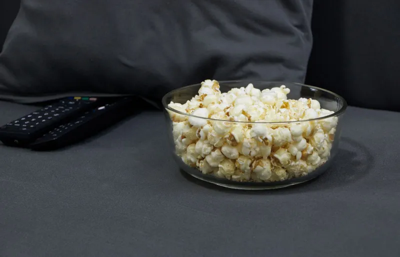A glass bowl of popcorn next to two TV remote controls on a grey sofa