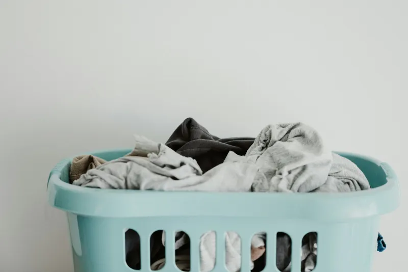 A pale blue plastic laundry basket full of grey and dark clothes against a white wall