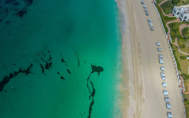 Aerial view of turquoise sea meeting a white sand beach lined with sun loungers and low resort buildings