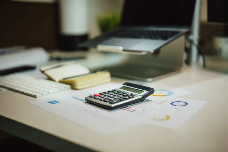 A calculator on printed charts beside a laptop on an office desk