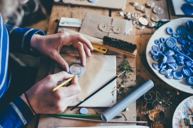 Hands working on a handmade craft project at a wooden table covered in supplies