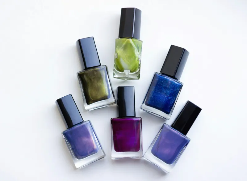 Six bottles of shimmering nail polish in green, blue and purple on a white surface