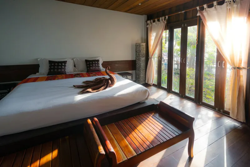 A sunlit hotel bedroom with a wooden bed, bench and tall windows