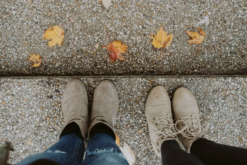 Two pairs of suede ankle boots on a pavement scattered with autumn leaves