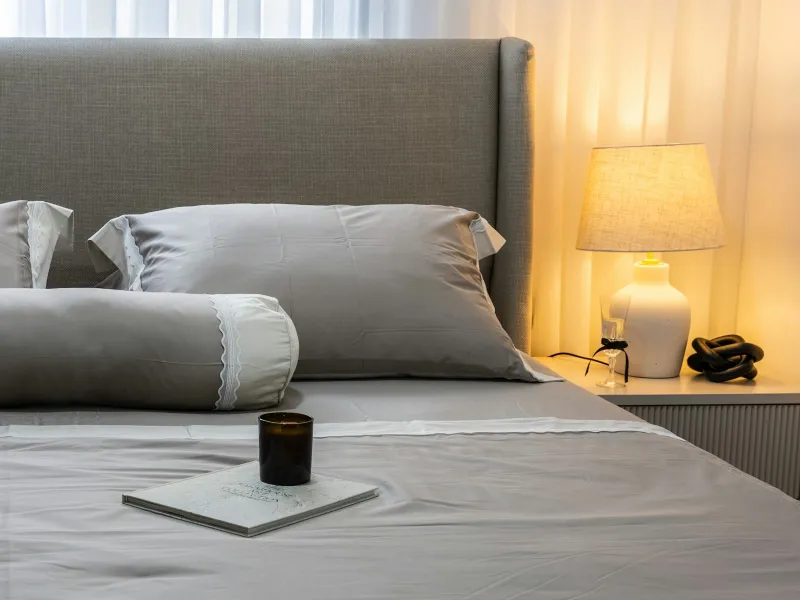 A neatly made bed with grey satin-look pillowcases beside a lit bedside lamp
