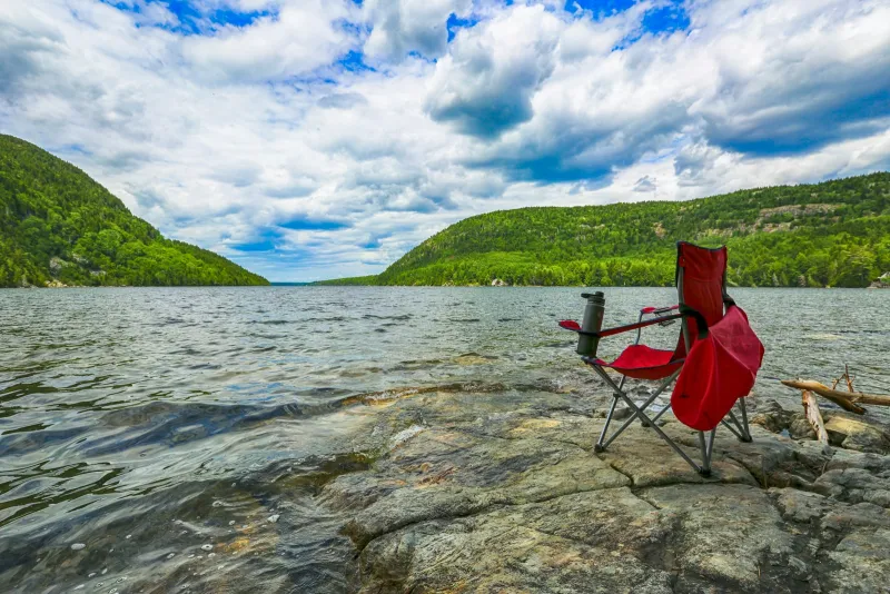 A red folding camping chair on a rocky lakeshore with wooded hills behind