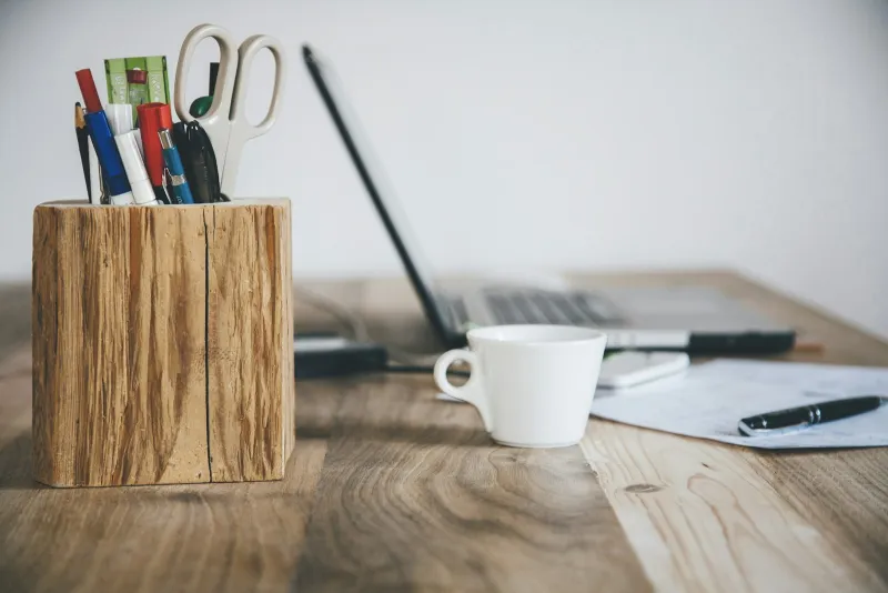 White cup on a wooden desk beside a pen pot and laptop
