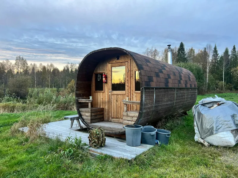 A wooden barrel sauna on decking in a grassy field at sunset