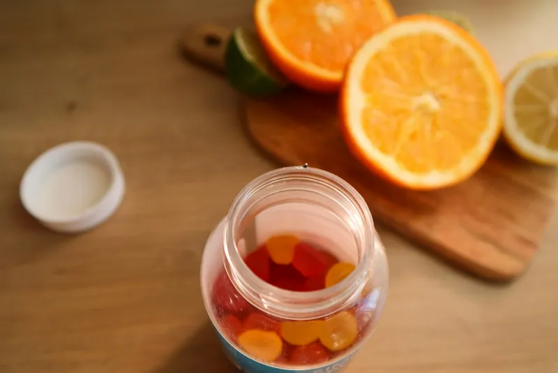An open jar of colourful gummies on a wooden table next to sliced oranges