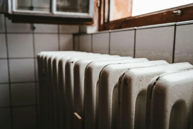A white column radiator under a wooden window on a tiled wall