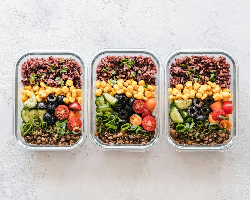 Three glass meal prep containers filled with rice, sweetcorn, olives, tomatoes and lentils