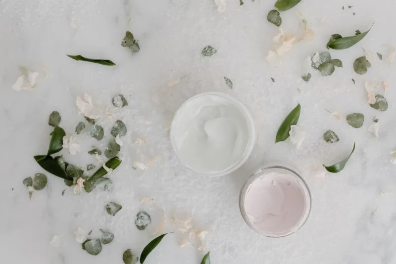 Open jars of body cream on marble scattered with leaves and petals