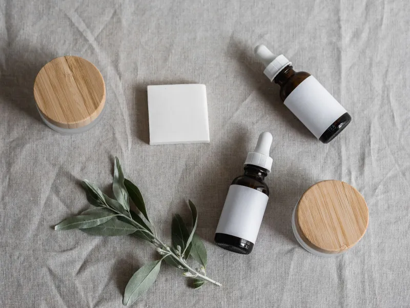 Unlabelled dropper bottles, bamboo-lidded jars, a soap bar and an olive sprig on linen